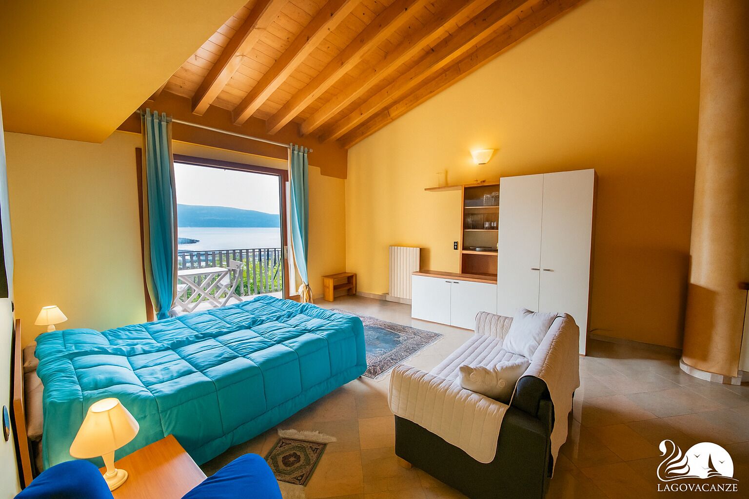 A bedroom with a large bed, a sofa, a desk, and a wardrobe. There is a window with a view of the sea.