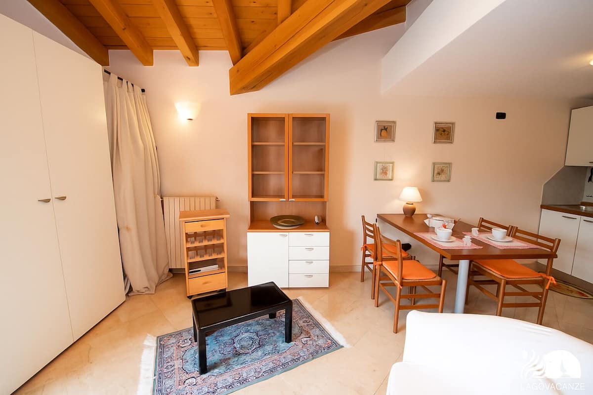 A compact room with a wooden ceiling, a dining table with chairs, a small kitchen area, and a wardrobe.