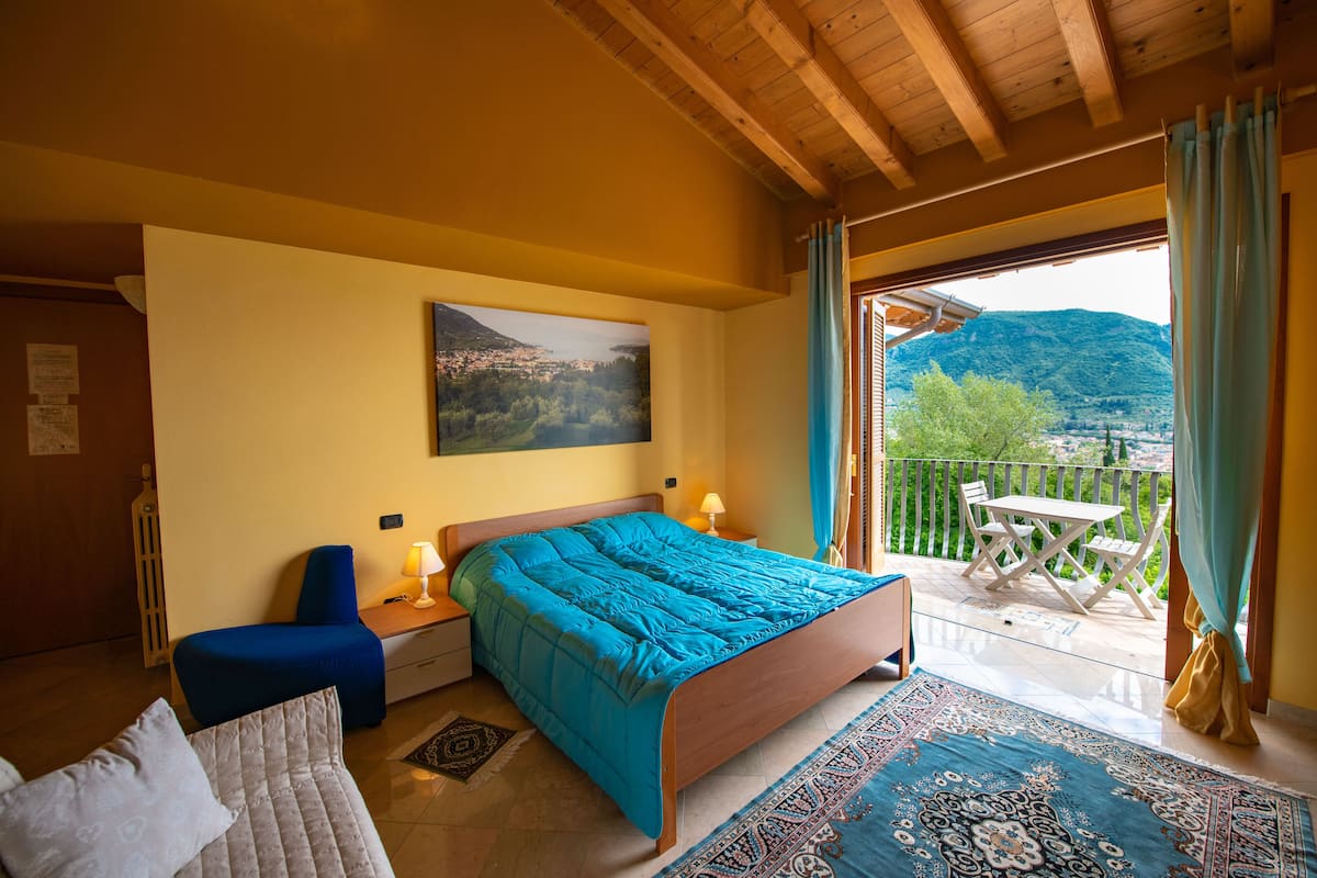 A bedroom with a bed, a chair, a nightstand, a table, and a balcony with a view of the mountains.
