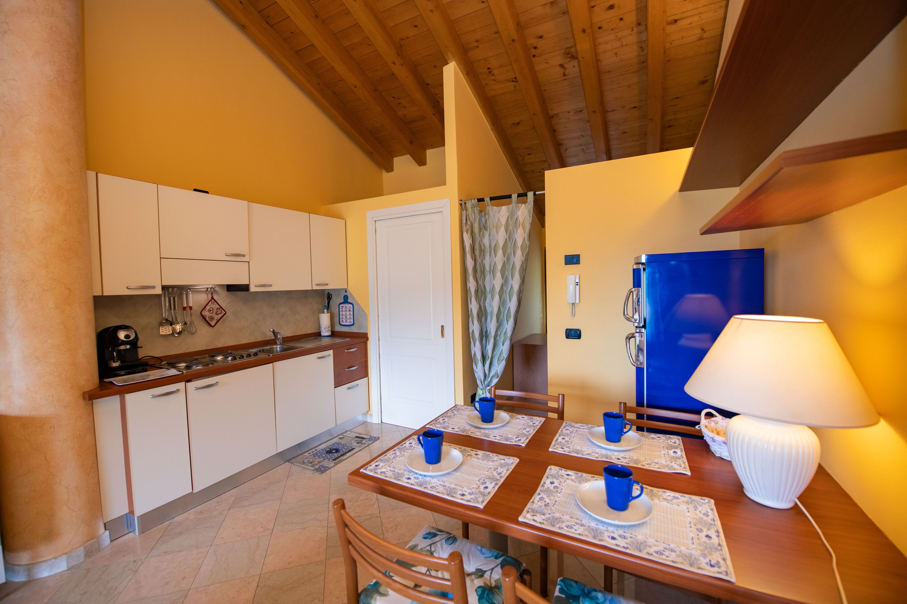 A compact kitchen with white cabinets, a blue refrigerator, and a dining table set with blue cups and plates.