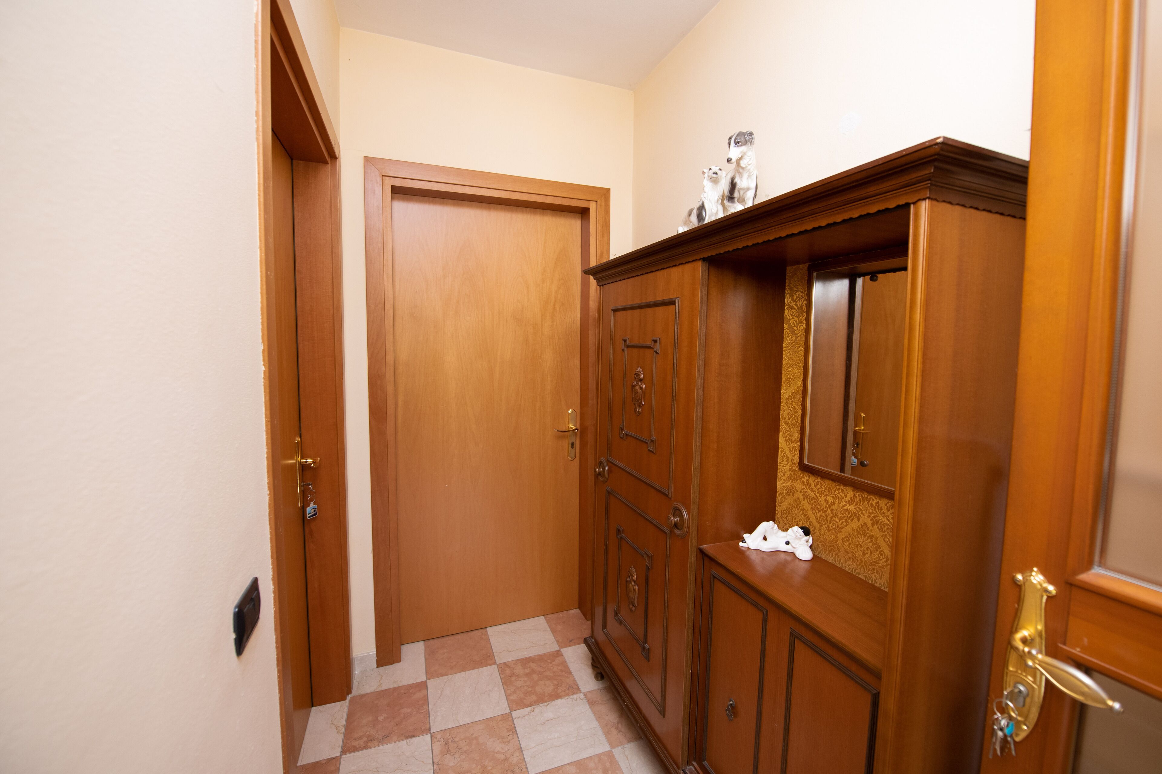 A hallway with a wooden door, a wooden cabinet with a mirror, and a tiled floor.