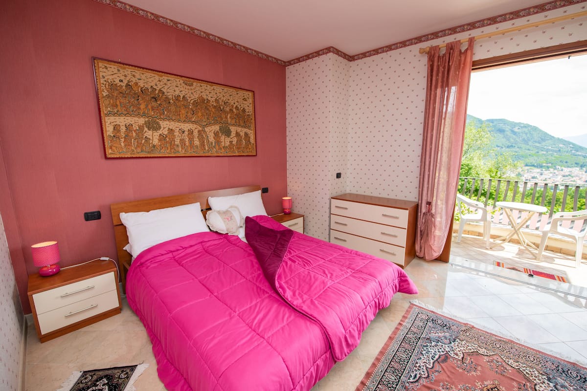 A bedroom with a large bed, a nightstand, a dresser, and a view of a balcony with outdoor furniture.
