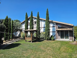 A two-story house with a wooden pergola in the front yard, surrounded by tall cypress trees.