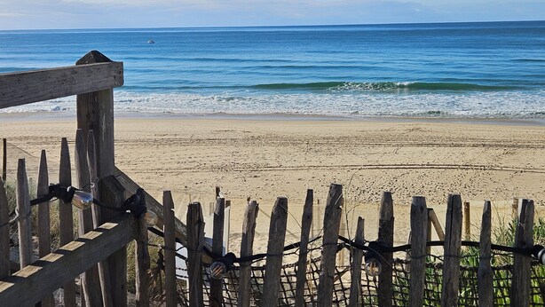 Une clôture en bois avec des barreaux métalliques, qui mène à une plage de sable fin aux vagues douces.