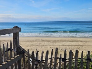 Een houten hek met metalen spijlen, dat uitkomt op een zandstrand met zachte golven.