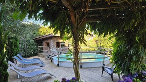 Une terrasse en bois avec un jacuzzi et des chaises longues, entourée de verdure et d’un petit bâtiment.