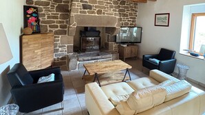 A cozy living room with a stone fireplace, wooden ceiling beams, a sofa, and a television. - Maison en Granit Typique Rénovée (Plounévez-Moëdec)