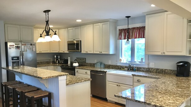 A spacious kitchen with granite countertops, white cabinetry, a central island with bar stools, stainless steel appliances, and a window with curtains. - Newly renovated, 4 bedroom, central location, Immaculate, bright Family Retreat! (edgartown)