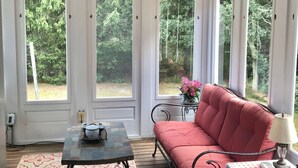 A sunlit room with a ceiling fan, a patterned rug, a red sofa, a small table with a lamp, and large windows with a view of greenery. - Newly renovated, 4 bedroom, central location, Immaculate, bright Family Retreat! (edgartown)