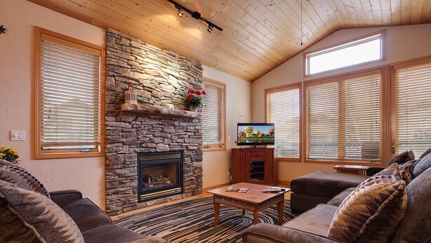 A cozy living room with a stone fireplace, wooden ceiling, and large windows with blinds.