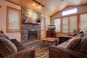 A cozy living room with a stone fireplace, wooden ceiling, and large windows with blinds.
