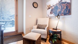 A cozy living room with a large window, a comfortable armchair, a small wooden table, a floor lamp, and a wall clock.