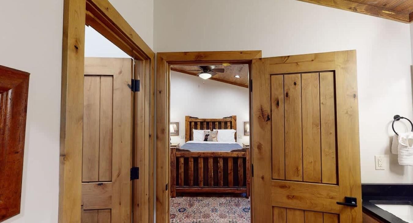 A wooden door leading to a bedroom with a bed, a ceiling fan, and a patterned rug.