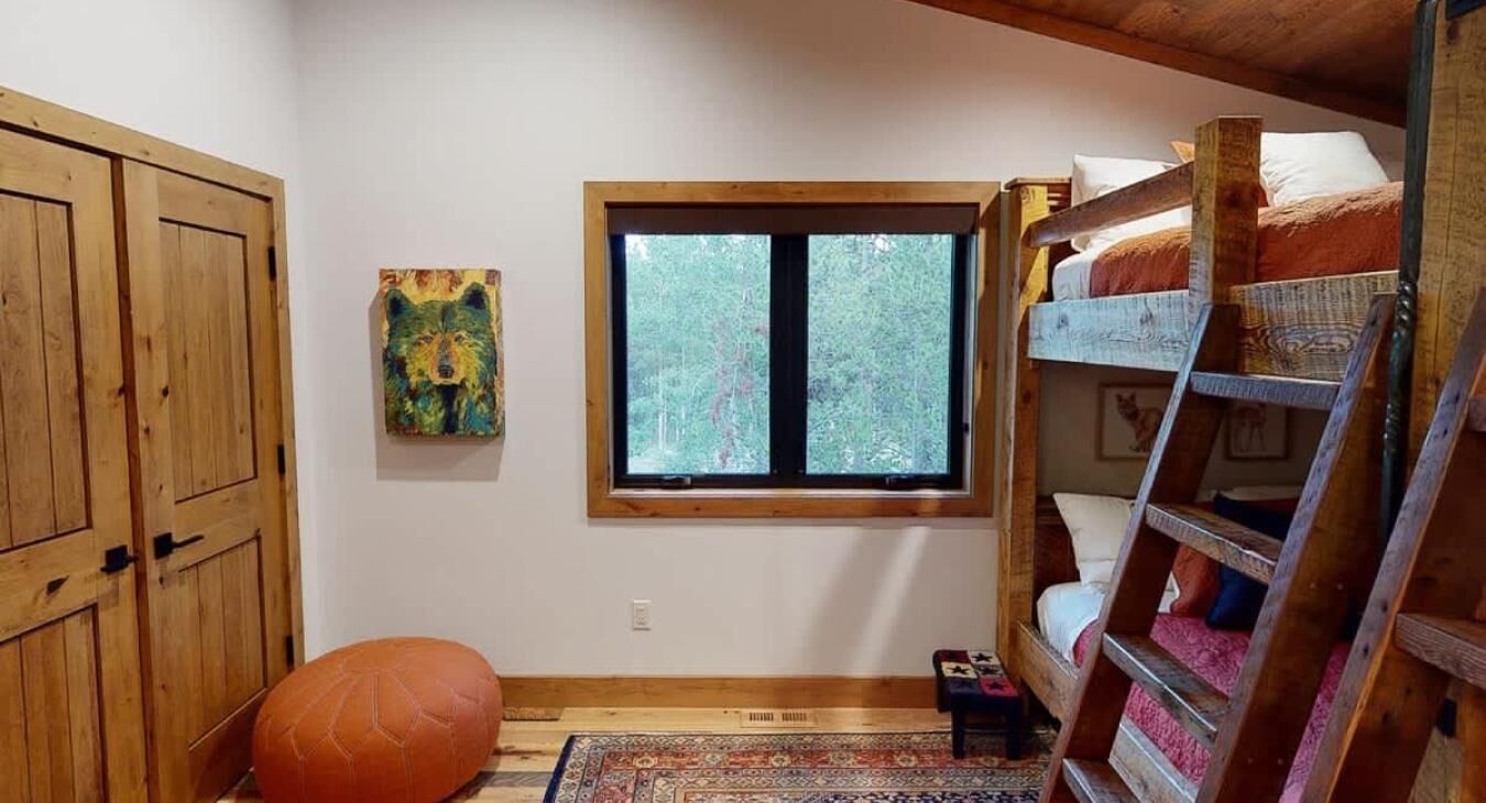 A room with a bunk bed, a window with a view of trees, a wooden wardrobe, and a colorful painting on the wall.