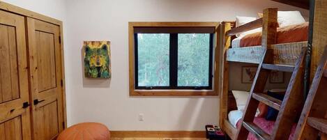 A room with a bunk bed, a window with a view of trees, a wooden wardrobe, and a colorful painting on the wall.