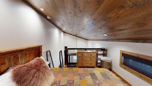 Ein Schlafzimmer mit schrägem Dach, Holz-Kopfteil, einem Bett mit flauschigem Kopfkissen, einem Schminktisch, einem Sessel, einem Fenster und einem an der Wand befestigten Fernseher.