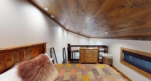Une chambre avec un plafond en pente, une tête de lit en bois, un lit avec un oreiller moelleux, une commode, une chaise, une fenêtre et un téléviseur fixé au mur.