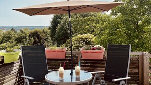 Una terraza de madera con dos sillas y una mesa bajo un paraguas grande, rodeada de plantas en macetas y árboles.