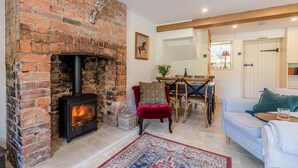 A cozy living room with a brick fireplace, a dining area with wooden furniture, and a blue patterned sofa. - Poppy Cottage - sleeps 4 guests  in 2 bedrooms (Broadwell)