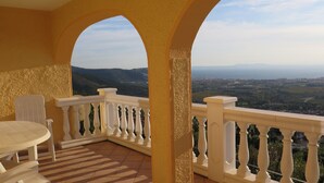 A balcony with white balustrades, a round table, and a chair, overlooking a scenic landscape with hills and a distant city. - lovely apartment with private swimmingpool and stunning views (Rosas / Roses)