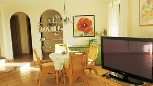 A living room with a dining table, wicker chairs, a flat-screen TV, a bookshelf, and a painting of a flower. - lovely apartment with private swimmingpool and stunning views (Rosas / Roses)