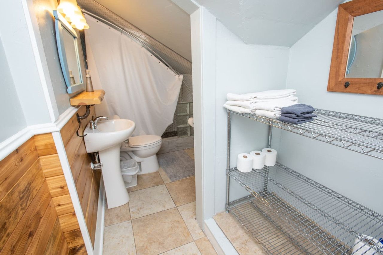 A compact bathroom with a toilet, sink, and shower area. There are shelves with towels and toilet paper.