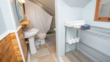 A compact bathroom with a toilet, sink, and shower area. There are shelves with towels and toilet paper.