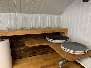 A kitchen with wooden shelves holding clear glasses, a roll of paper towels, and stacked plates.