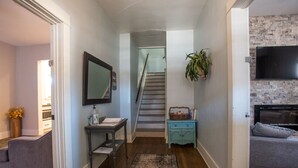 Un pasillo con una escalera, una pequeña mesa con un florero, un sofá gris y un televisor montado en una pared de ladrillo.