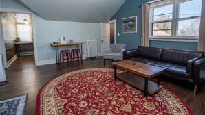 Un salon avec un canapé en cuir foncé, une table basse en bois et un tapis à motifs rouges. On y trouve une fenêtre avec des rideaux beige, un radiateur blanc et une petite table accompagnée de deux tabourets.