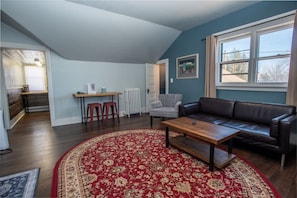 A living room with a dark leather sofa, a wooden coffee table, and a red patterned rug. There is a window with beige curtains, a white radiator, and a small table with two stools.