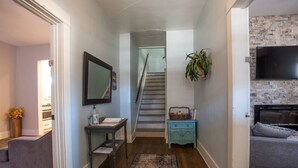 Un couloir avec un escalier, une petite table sur laquelle est posé un vase, un canapé gris et un téléviseur fixé à un mur en briques.