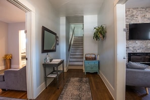 A hallway with a staircase, a small table with a vase, a grey sofa, and a TV mounted on a brick wall.