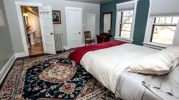 Une chambre avec un grand lit, un tapis à motifs, une porte donnant sur une salle de bain et une fenêtre avec des stores.