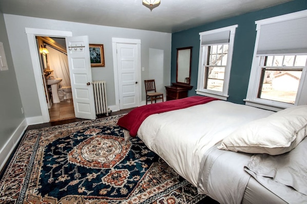A bedroom with a large bed, a patterned rug, a door leading to a bathroom, and a window with blinds.