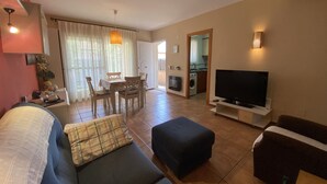 Ein Wohnzimmer mit Sofa, Hocker, Fernseher und einem Essbereich mit Stühlen und Tisch.