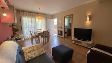 Een woonkamer met een bank, een poef, een televisie en een eethoek met stoelen en een tafel.