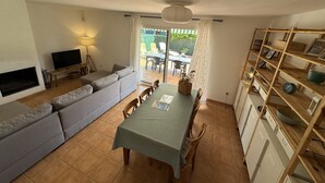 A living room with a sofa, TV, and dining table. There is a patio with chairs and a table outside. - Daró 3D 49 - Aircon, pool, corner house, 250m from the beach (Torroella de Montgri)