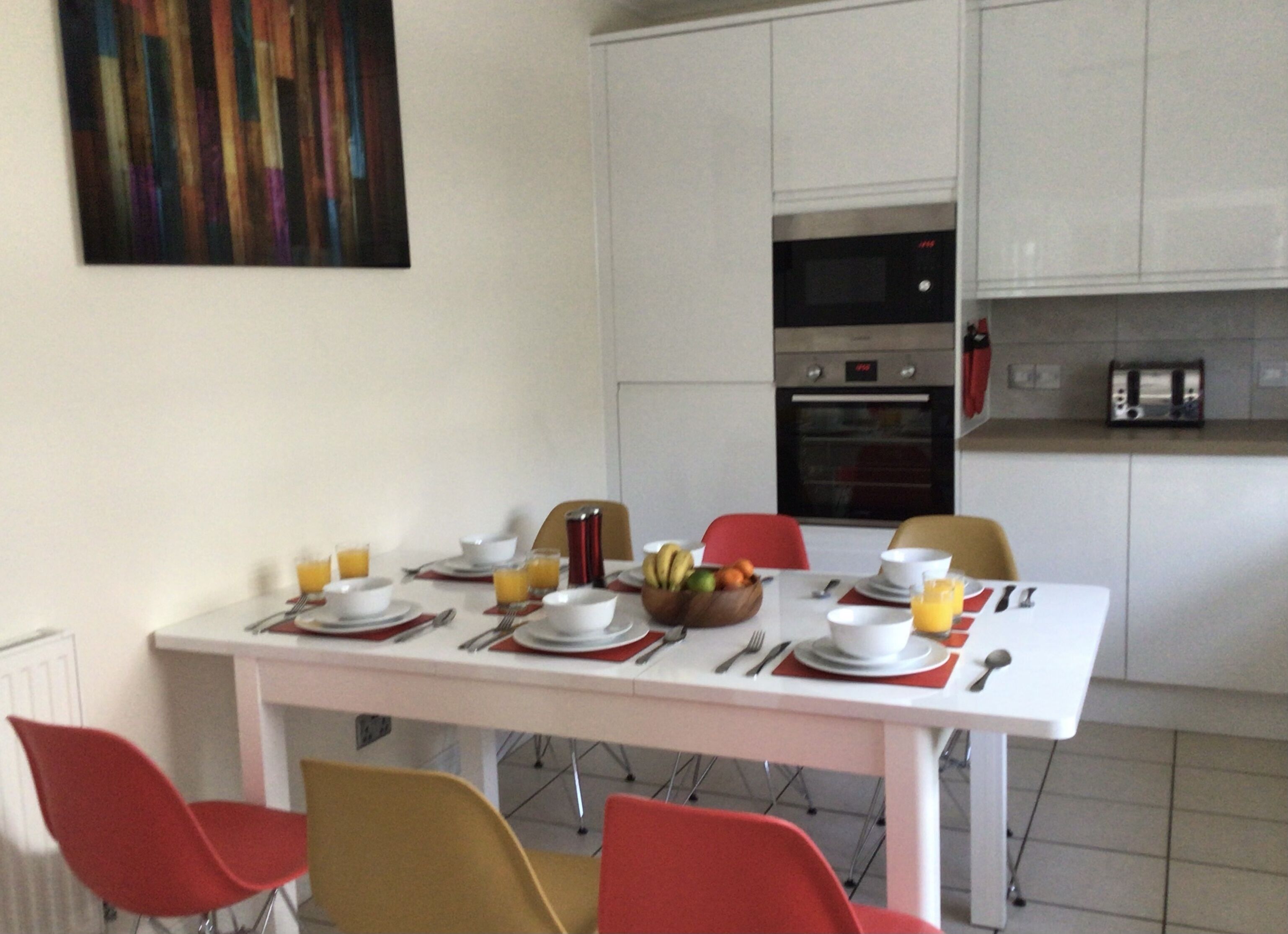 A dining area with a white table set for four, red chairs, a fruit bowl, and a red and white patterned table runner.