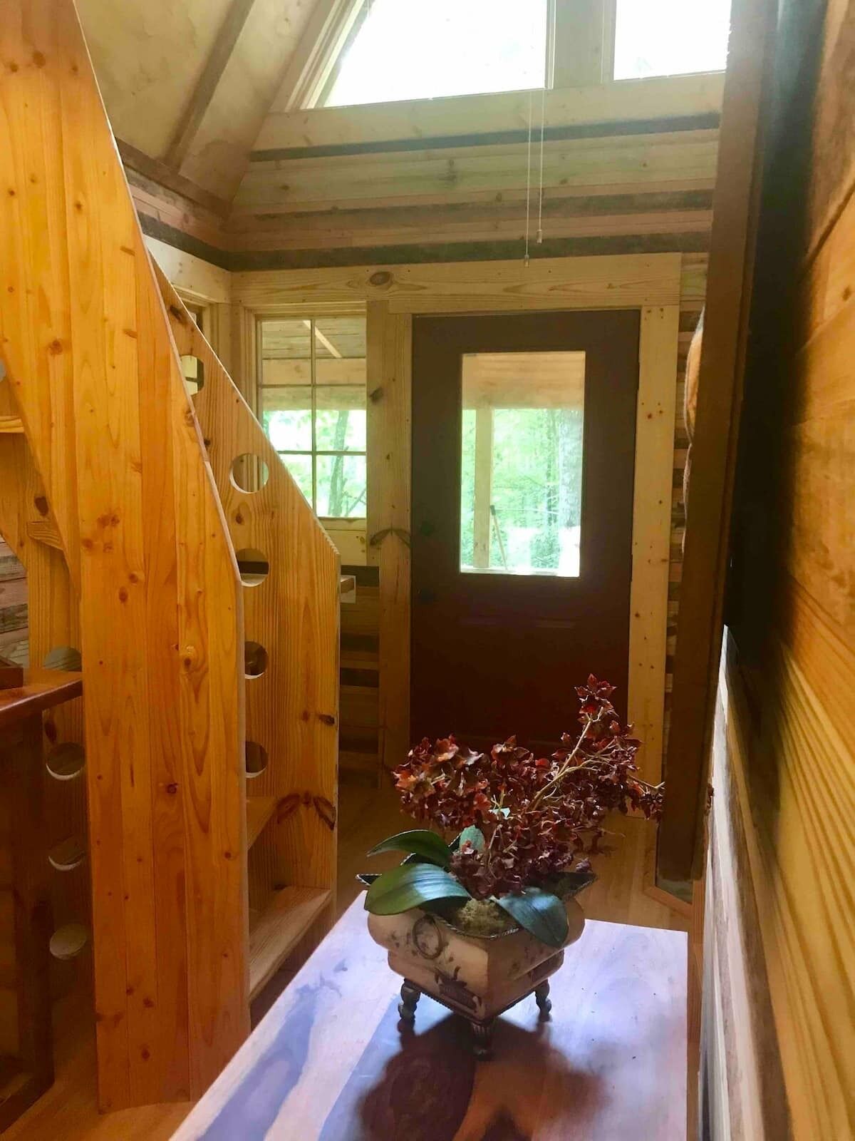 A wooden interior with a staircase, a door leading outside, and a vintage-style table with a vase of flowers.