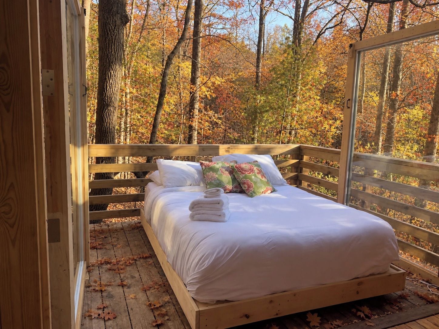 Ein hölzernes Bett mit weißer Bettwäsche und blumigen Kissen, vor einem Hintergrund aus Herbstlaub.