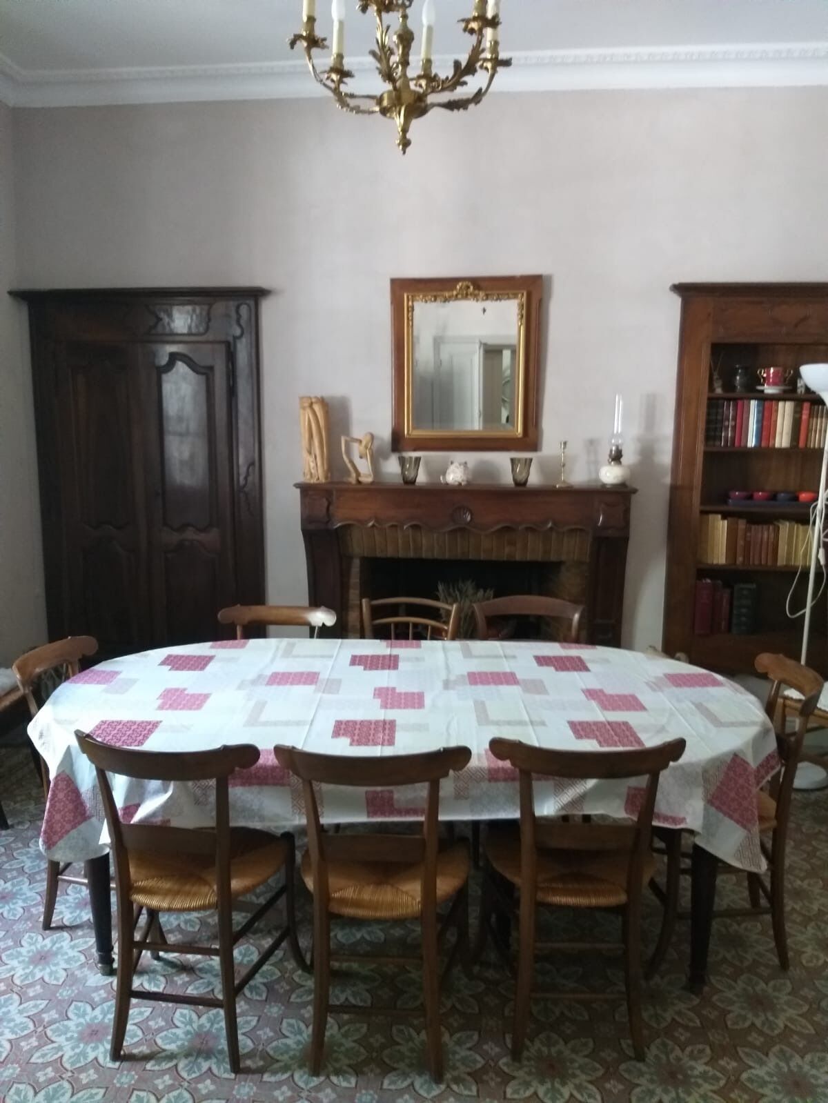 A dining room with a round table covered in a white tablecloth with pink patterns, surrounded by wooden chairs. There is a chandelier above the table, a wooden cabinet to the left, and a bookshelf to the right.
