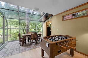 A room with a foosball table, a dining table with chairs, and large windows offering a view of trees.