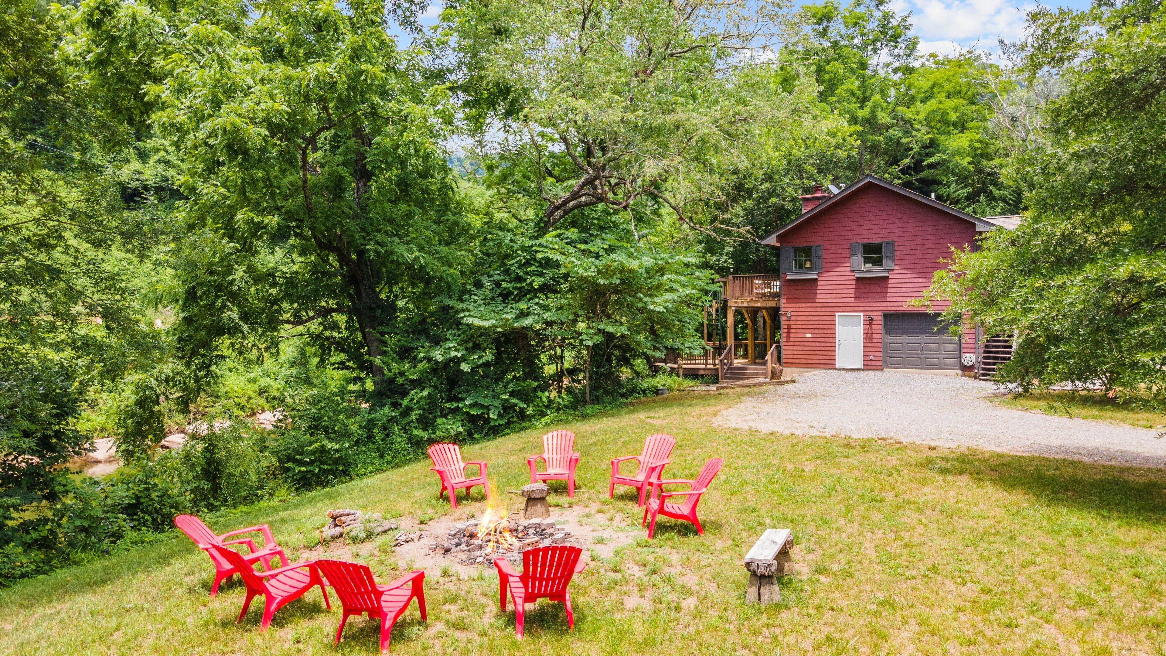 A red cabin with a deck, surrounded by lush greenery and a fire pit with red chairs.