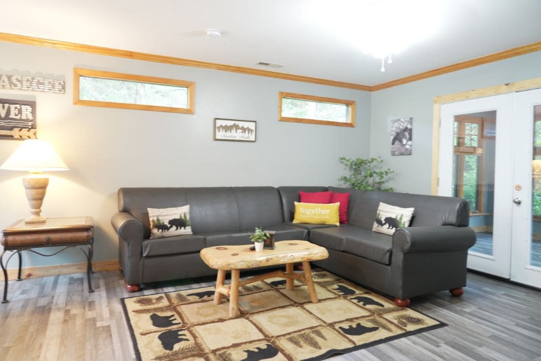 A living room with a grey sofa, a wooden coffee table, and a patterned rug. There are framed pictures on the wall and a lamp on a side table.