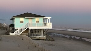 Ein zweistöckiges Strandhaus mit Balkon, gelegen an einem sandigen Strand mit dem Meer im Hintergrund.
