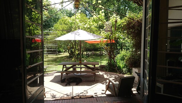 Een open terras met een houten tafel en bankjes, omringd door weelderige begroeiing en een hangende vogelkooi.