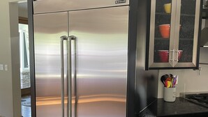 Frigorifero in acciaio inox con congelatore integrato, un piano di lavoro nero e un pensile con vetro, contenente contenitori rossi e gialli.