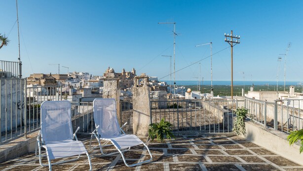 Una terraza en la azotea con dos sillones blancos, vistas a un paisaje urbano y un cielo azul despejado.