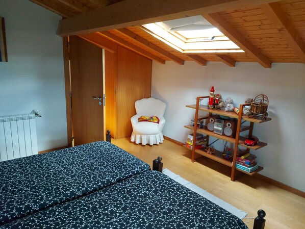 A bedroom with a skylight, a white chair, a bed with patterned bedding, and a wooden shelf with toys and books.